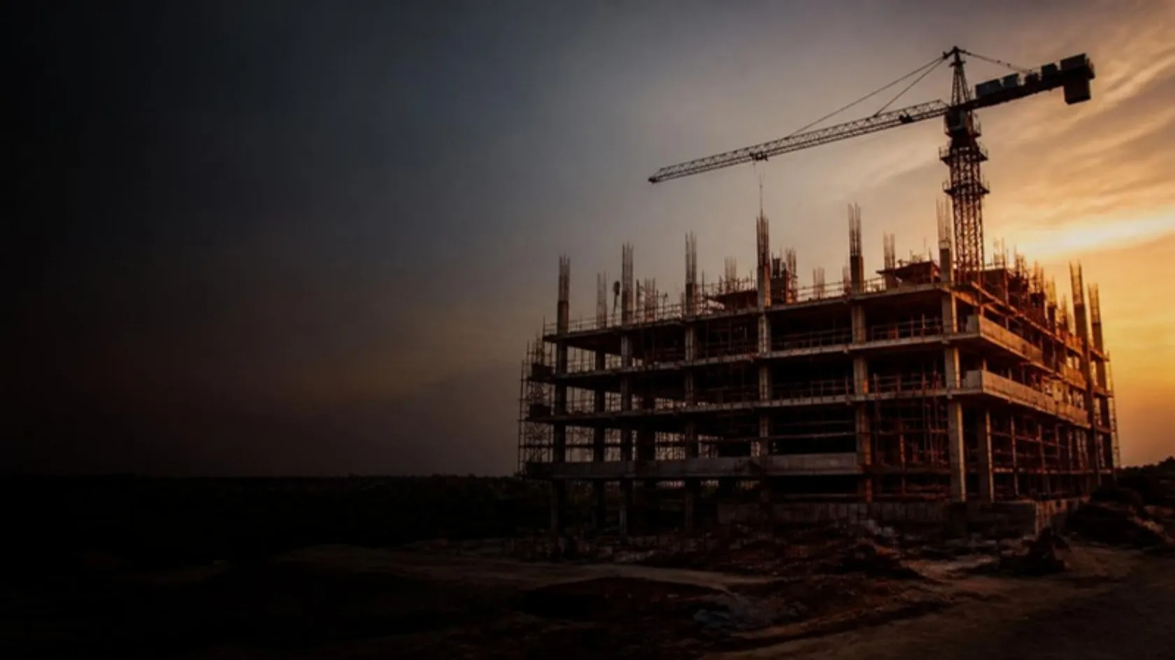 A multi-storey concrete building under construction with a tower crane at dusk
